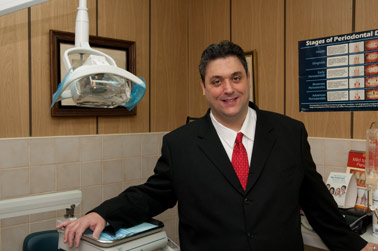 A man wearing a suit stands in front of a dental office setting with a sign and medical equipment visible.