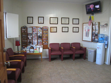 The image shows an interior view of a waiting room with chairs, a television screen displaying news, and framed diplomas on the wall.