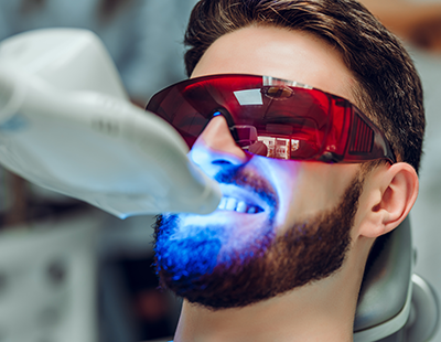 A person with blue light emanating from their mouth, wearing red goggles and a white dental mask, appears to be smiling while seated in a dental chair.
