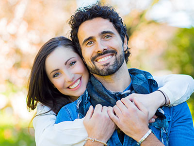 The image shows a man and woman embracing each other in an outdoor setting, both smiling and appearing happy.