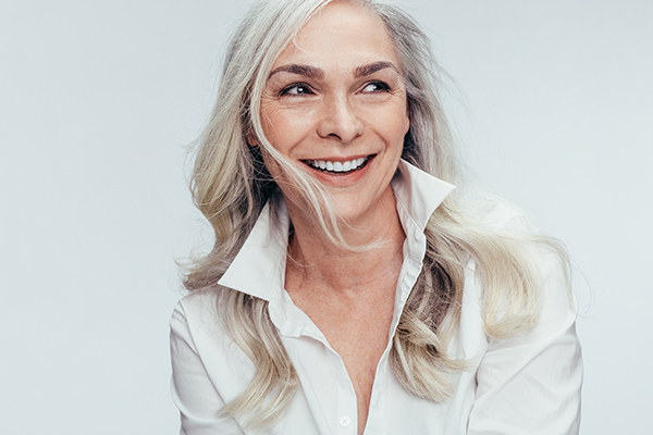 The image features a woman with short blonde hair smiling at the camera. She has her left hand on her face, holding up her cheek, and appears to be wearing makeup. Her attire includes a white top. The background is plain and light-colored, providing a clear contrast to the subject.