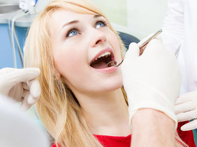The image shows a woman sitting in a dental chair with her mouth open, receiving dental care from a professional who is using dental tools and wearing gloves and protective eyewear.