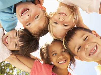 The image displays a group of children posing together with smiles, under a bright sky, likely taken during a sunny day outdoors.