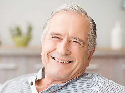 The image shows an elderly man with gray hair, smiling at the camera while sitting comfortably indoors, wearing a blue shirt and glasses, with a relaxed posture.