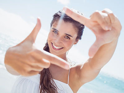 A young woman with long hair is smiling at the camera while holding up her hand to frame the shot.