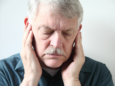 The image shows an older man with his eyes closed, holding his face with both hands, appearing to be in pain or discomfort.